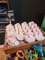 A photo of a square box containing  a bunch of white ghost shaped bath bombs with a rainbow on the side of them on a light brown table with a black card next to them stating ghostie bath bomb in white font on a bright background