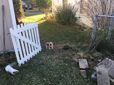 A white picket gate leaning slightly against a white-trimmed tan house corner, leaving a wide gap between itself and the corner of a chainlink fence. A small tan stone block with the University of Nebraska N and "Huskers" writtenacross teh center stands in the middle of the gap.