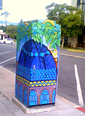 A utility box painted with a building that has a blue dome.  There is lots of Big green leaves as well.  In the background of the picture is a palm tree.