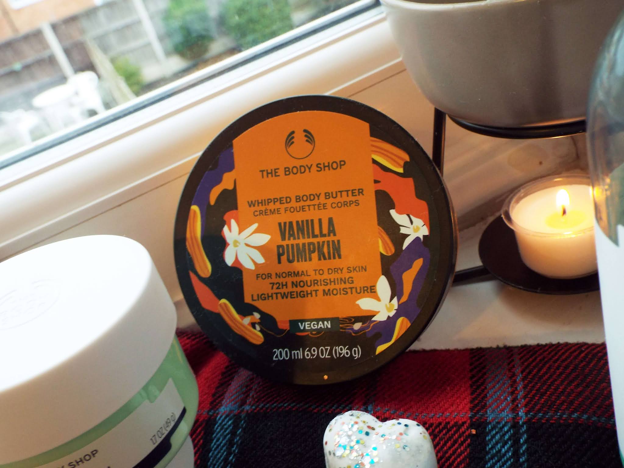 The Body Shop Vanilla Pumpkin Body Butter, sat on tartan scarf in the nook of a windowsill, with wax melter burning to background and other surrounding beauty products.
