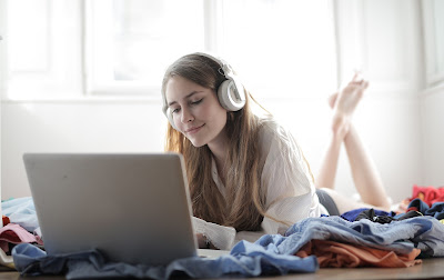 young girl listening to a podcast with earbuds on her laptop while laying on a bed.