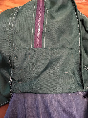 A roughly rectangular piece of sark hunter green fabric, with the edges folded under and pinned over the end of a navy zipper in the side of a dark hunter green bag with white stitching.
