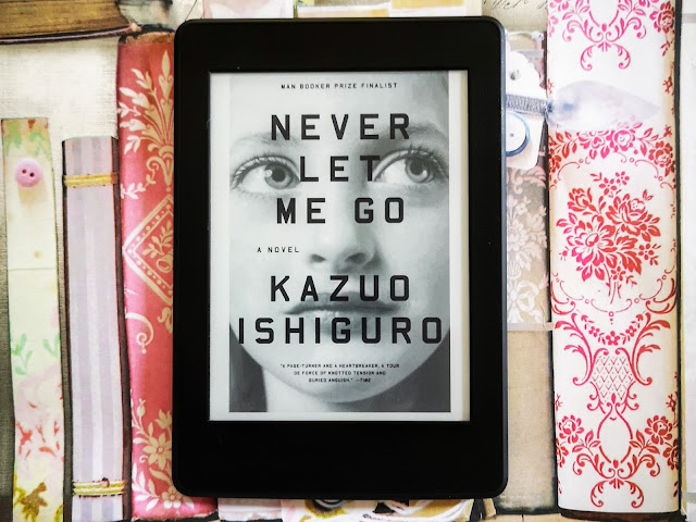 Image of the front cover of the ebook version of Kazuo Ishiguro's novel Never Let Me Go on a Kindle Paperwhite. The cover features and image of a close up of an adolescent girl looking upwards. The Kindle is placed on a floral background.