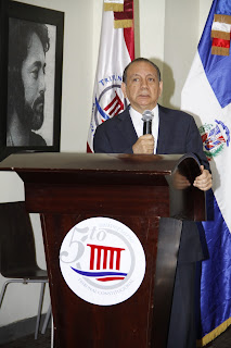 El Dr. Diómedes Núñez Polanco, director de la BNPHU se dirige al público presente al inicio del Taller desde el pódium. Viste traje azul marino, camisa blanca, corbata de rayas en diagonal azules, grises y negras. Cara redonda, expresión seria, tez clara, pelo negro con entradas, frente amplia, cejas desiguales, ojos claros, nariz ancha, boca de labios finos, orejas grandes, cuello corto. Sostiene el micrófono con la mano derecha mientras con la izquierda agarra el borde del pódium. El pódium es de madera color caoba y tiene en la parte frontal el logos del Tribunal Constitucional. A su espalda las banderas: de izquierda a derecha, la bandera del Tribunal Constitucional, color blanco y rojo, con letras y símbolos azules; la bandera nacional dominicana, sus dos cuartos azules y el blanco de la cruz. En la pared izquierda, en blanco y negro, una foto ampliada de un escritor dominicano.