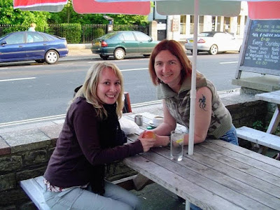 two women sitting outside a pub, one is pretty and blond, one is me, dark haired and wearing a camo jacket with no sleeves, the tattoo is visible
