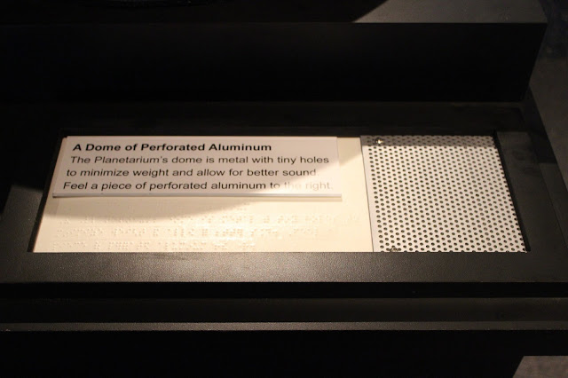 In large print and Braille reads the following exhibit signage: "A Dome of Perforated Aluminum.  The Planetarium's dome is metal with tiny holes to minimize weight and allow for better sound.  Feel a piece of perforated aluminum to the right."  To the right of the text is a piece of silver aluminum with many small holes in it for visitors to feel.