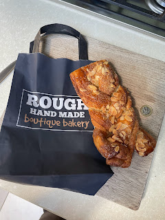 A small black paper bag with Rough Hand Made Boutique Bakery is lying on the kitchen counter. A large plaited pastry is on top, with a £1 coin next to that. The pastry is big.