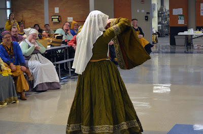 A young woman with her back to the camera, wearing a white veil and a flowing, green-gold medieval gown with trailing sleeves, all trimmed in gold vining oak leaves. She's holding her right elbow high to show the lacing at her sides, through which you can see her white chemise, and her narrow red and gold belt sits at her waist.