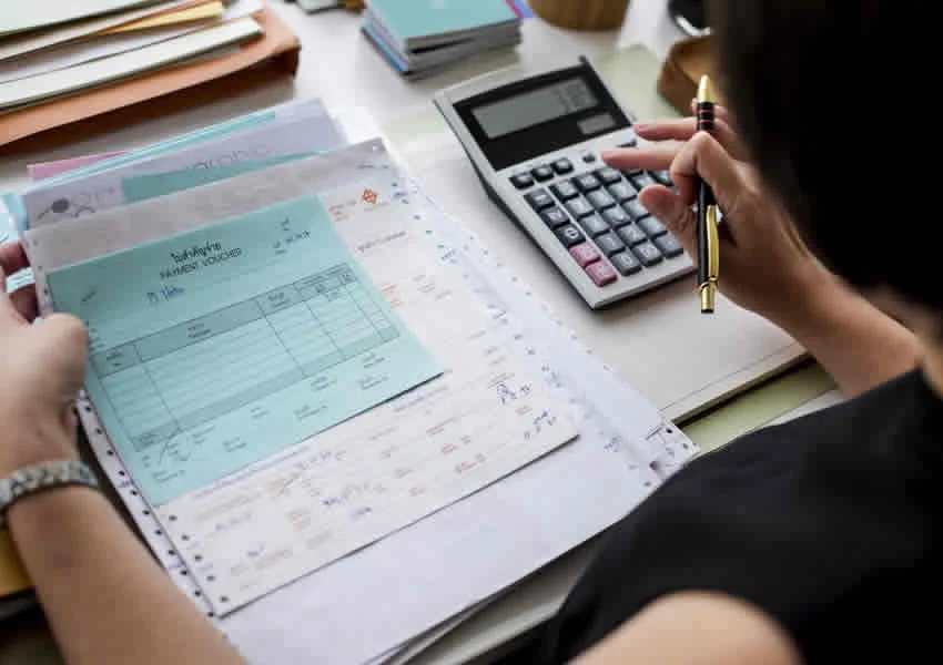 Imagem mostra uma pessoa em uma mesa digitando em uma calculadora e segurando muitos boletos com a outra mão.
