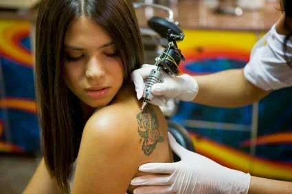 Vemos a un tatuador tatuando a una chica jovencita muy guapa, ella pone cara de dolor mientras observa al tatuador,