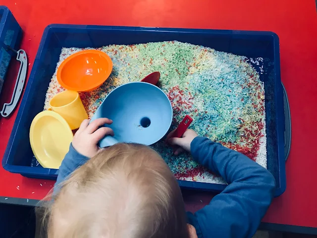 A toddler sitting in front of a tray of coloured rice with a spoon, bowl and funnels