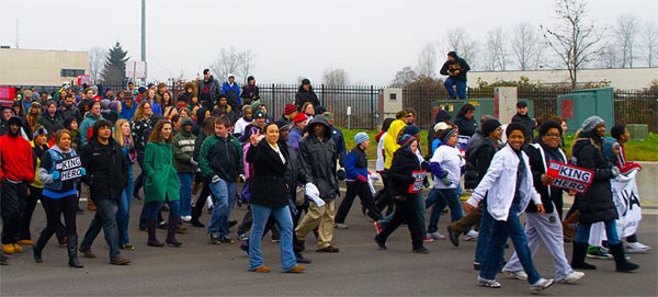 a crowd of people walks by, some carrying signs that read "KING HERO"