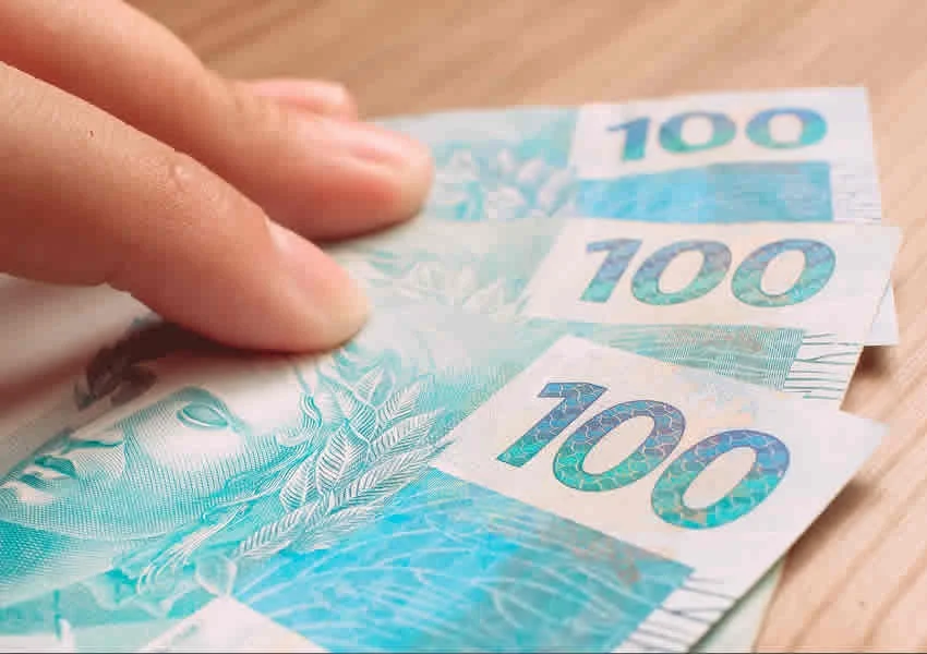 Mão sobre uma mesa de madeira segurando algumas notas de 100 reais.