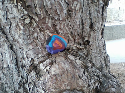 Small rock painted multiple colors, resting on a knot in a tree trunk.