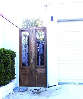 A timber door, weather-stained and with some iron work in place of what might have been the top panel, set into a wall surrounding a house. Its clear that there's a garden beyond, but you can't really see it (without getting closer and peering in, which would be rude!).