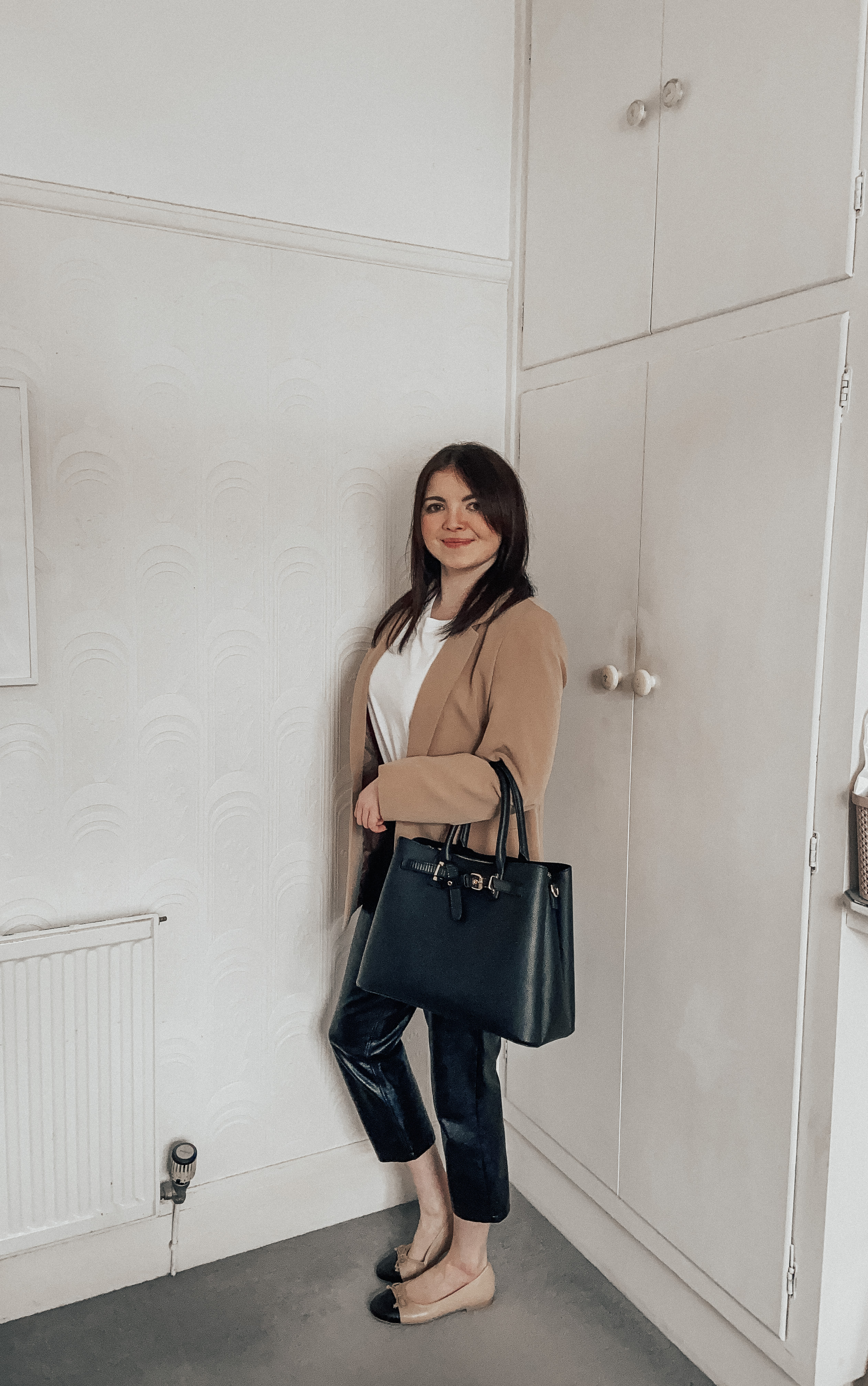 A woman wearing a beige blazer, white tee, leather cropped trousers and Chanel flat dupes.