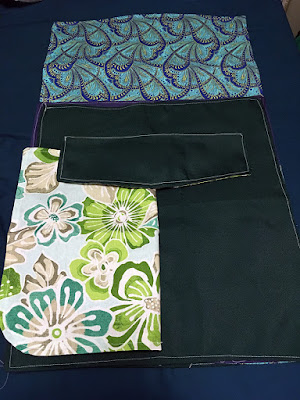 Several pieces of white-stitched dark hunter green, white, teal, and green tropical floral, and blue and navy art deco fabric, laid out to form the outline of a large flat envelope with open flap, and a smaller flap centered high on the larger envelope, with the white, teal, and green tropical floral fabric arranged as a large pocket under half of the small flap.