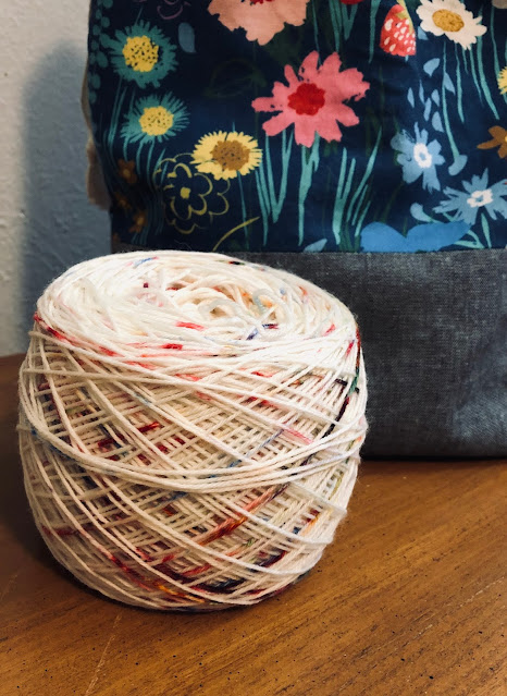 A ball of yarn sitting on a brown hope chest. The yarn is white based with speckles of multiple colors. There is a project bag in the back that has a gray bottom and a blue accent piece with flowers and strawberries.