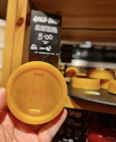 A photo of a white hand holding a circular gold coin shaped piece of soap on a bright background with a light brown shelf and black rectangular card that says gold coin soap lush in white font on a bright background