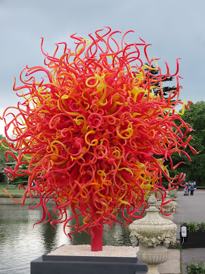 photo of a round, tree shaped  Dale Chihuly glass sculpture - predominately  red , standing in front of a lake