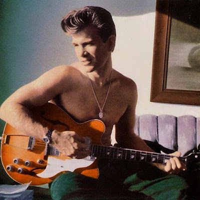 Chris Isaak color photo sitting shirtless on a bed, playing a guitar wearing green pants