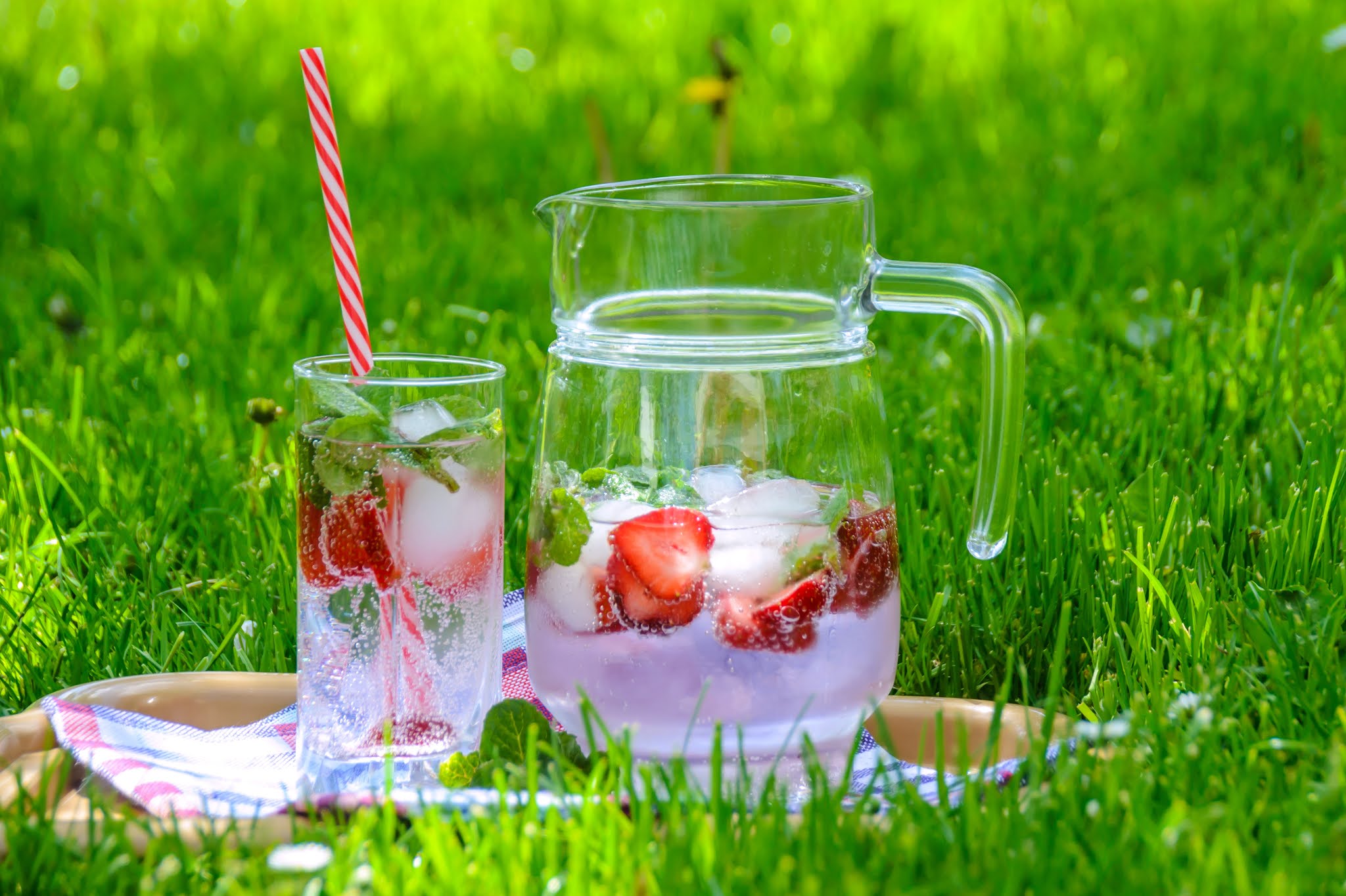 ice water with cut up strawberries in pitcher with glass laying on grass