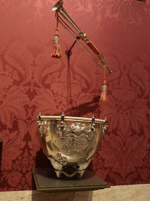 Photo. A ceremonial Baroque trumpet from Germany, very long, with yellow and gold tassels hanging from both ends, in front of a wall with red brocade wall paper. Below the trumpet is a baroque kettledrum, made of copper