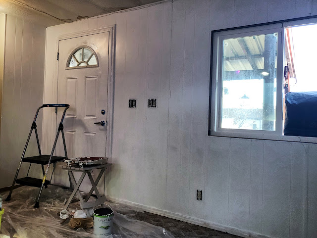 A room is mid-renovation with walls partially painted in light color, displaying a stepladder, paint tray, and an open paint can on the floor. Clear plastic sheeting is laid down to protect the floor, and the window is partially covered, implying ongoing work.