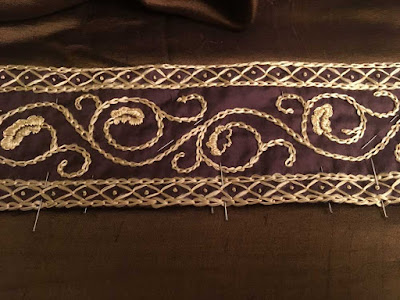 A close-up of a band of gold oak-leaf embroidery with narrow diamond borders, pinned at the lower edge to a piece of green-gold silk.