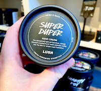 A photo of a white hand holding a black cylindrical tub filled with yellow liquid with a black lid that says super duper hand lotion lush in white font on a bright background