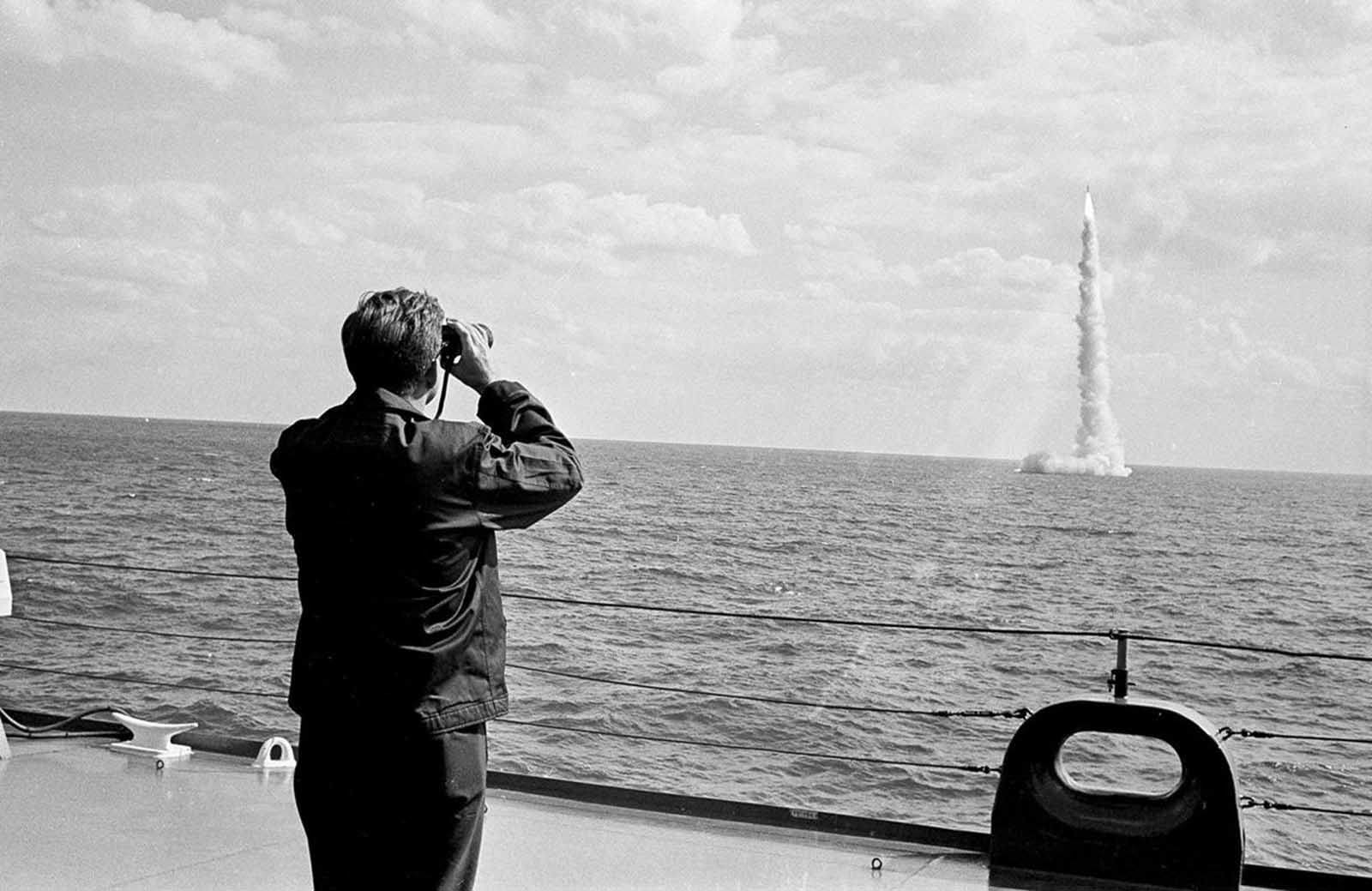 President John F. Kennedy, wearing a windbreaker, watches through binoculars as a Polaris missile clears the surface after firing from a submerged nuclear sub off Cape Canaveral, Florida, on November 16, 1963. 