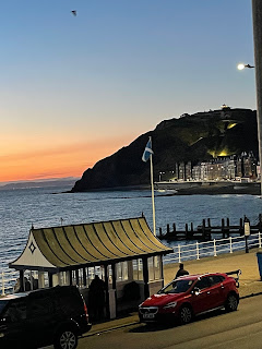 A photo of a beach. The sun is setting and the sky is orange and blue. On the right there's a cliff.