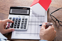 A hand using a calculator, which is on a desk surrounded by papers and a small model of a house.