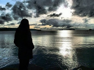 Una mujer de espaldas, mira al mar, con un cielo cubierto de nubes