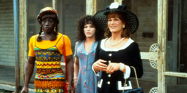 A film still from To Wong Foo. Three drag queens stood in front of a house. One of them is smiling.