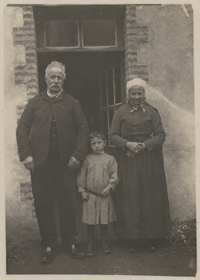 Un couple âgé se tient devant une porte ouverte, l'homme est moustachu et souriant, la femme porte une petite coiffe paysanne. Entre eux deux, un petit garçon en blouse rayée se tient droit.