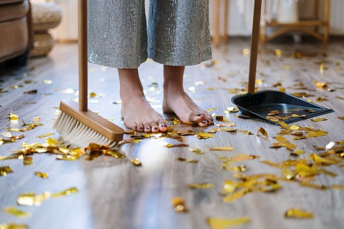 Pessoa descalça usando calça de paetês com vassoura e pá juntando serpentina dourada espalhada no chão da casa