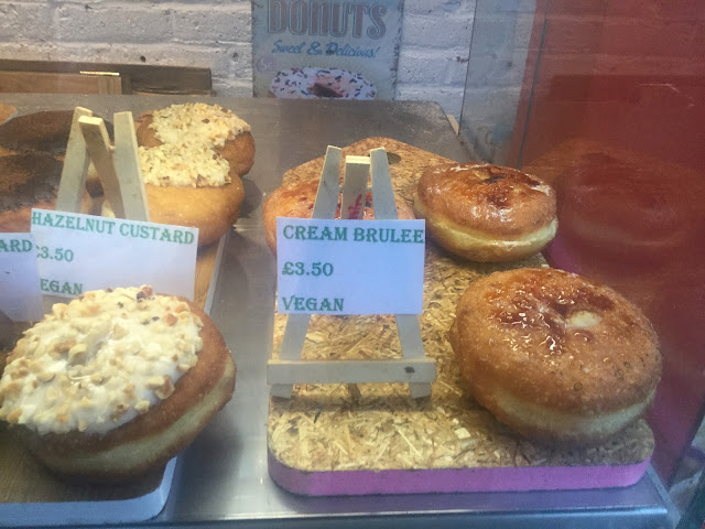 A donut display featuring round filled donuts l with a cream and chopped nut topping labelled as Hazelnut custard vegan £3.50. Alongside  another display of donuts which are labelled  Cream Brûlée Vegan £3.50