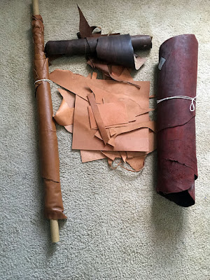 Three rolls of leather in an open-bottomed square, with scraps of pale vegetable-tanned leather in the center. The left edge is a narrow roll of caramel-brown on a cardboard tube; the top is a short, ragged-edged roll of dark brown; and the right side is a large, string-tied roll of deepest oxblood red.