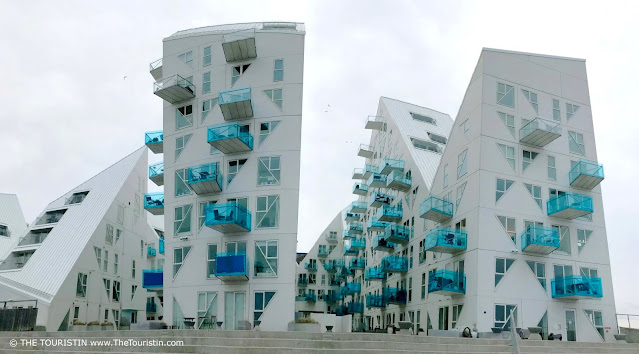 Several white multi-storey high rises in the form of icebergs with see-through light blue glass balconies on their facades, under a grey sky.