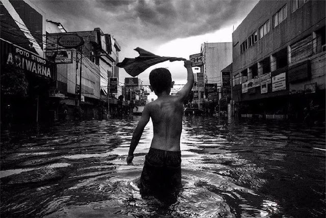Um menino gira sua camisa acima de sua cabeça em primeiro plano. Ele está imerso até os joelhos numa rua inundada em Jakarta. Ao seu redor, diversos estabelecimentos comerciais se estendem numa foto em preto em branco destacando o céu carregado.