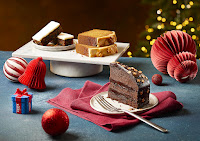 A wide angled photo with a dark brown rectangular table featuring some circular white plates filled with a slice of dark brown tall chocolate cake, a slice of rectangular light brown toffee loaf cake and a rectangular Christmas cake slice with white icing on a bright background