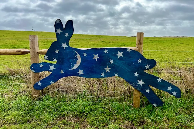 A wooden 2d hare on a fence painted blue with a moon and stars on