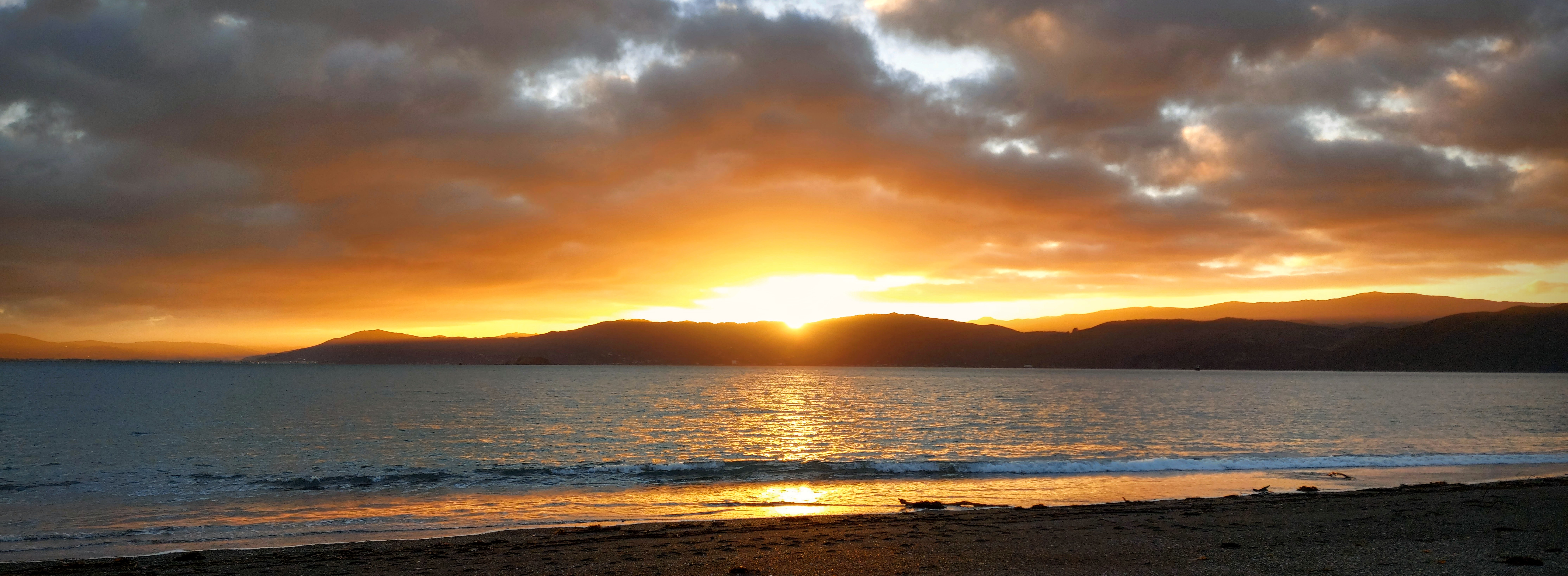 Sunrise over the distant Wellington harbour hills as seen from Seaton Beach, lighting up the clouds with amber and reflecting in the calm waters