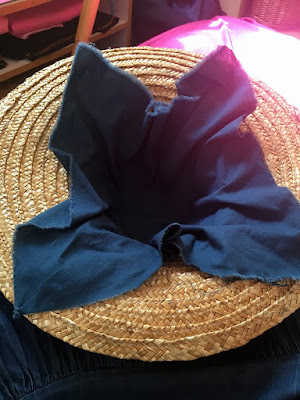 An upside-down straw hat, with a square of medium-blue cotton fabric roughly pushed into the crown, leaving the frayed edges and corners to stick out over the brim. Brilliant sunlight glows off the fuchsia sheets in the background, gleaming pinkly through one upturned corner of blue cloth.