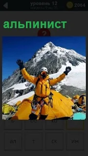 Около палатки на фоне вершины стоит альпинист с раскинутыми руками тепло одетый