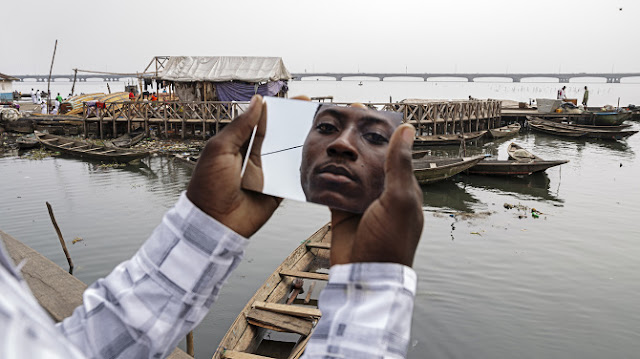 Braços seguram um espelho que enquadra o rosto de um homem negro que encara fixamente o observador. Ao fundo da foto se encontra uma construção ribeirinha e várias canoas no mar.