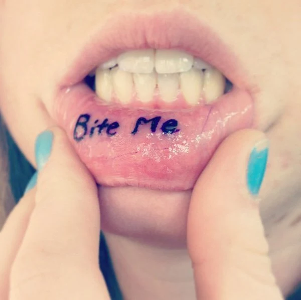 chica con las uñas pintadas de azul nos enseña tatuaje en el labio de la palabra muerdeme