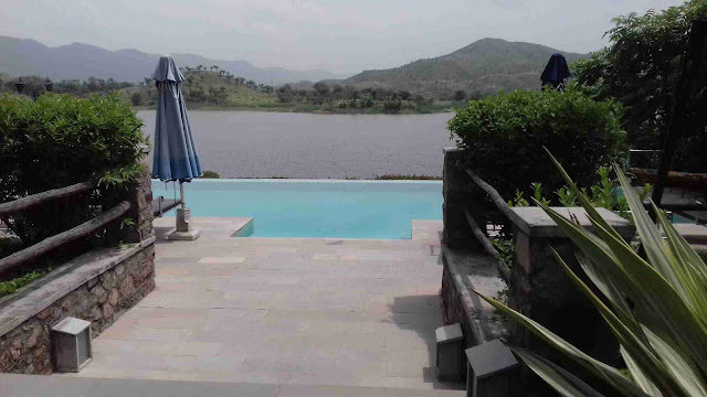 "A rectangular swimming pool with blue water, surrounded by lush greenery. A lake and mountains can be seen in the distance."