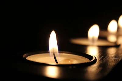 Uma chama de vela em primeiro plano, na escuridão atrás dela, outras chamas. #PraCegoVer #ParaTodosVerem